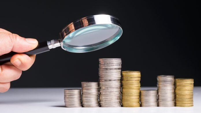 Man holding magnifying glass above stacks of coins.