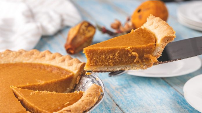 Closeup perspective shot of a freshly cut slice of pumpkin pie being lifted out of a pie tin on a pie server with plates and objects in the background on a blue painted rustic wooden table surface.