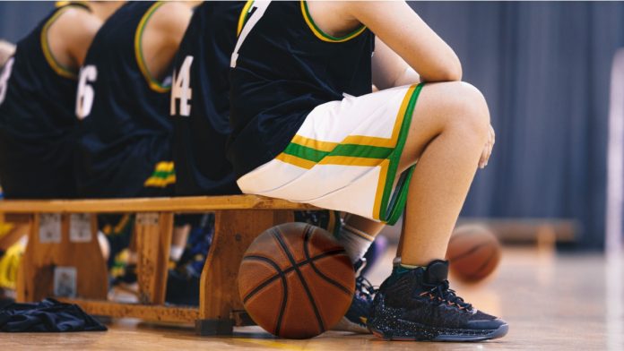Basketball Players Sitting on Wooden Bench. Group of Teenage School Boys Watching Basketball Match on the Court. Kids in Basketball Uniforms as Substitute Players.