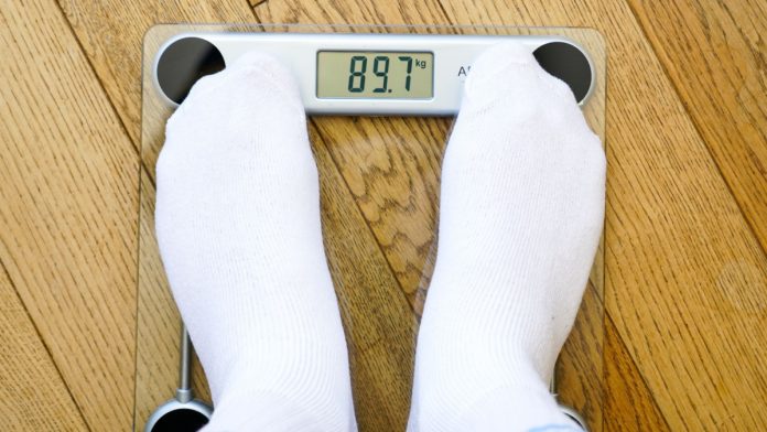 A man in white socks weighing himself on a transparent electronic floor scale, the weight reading is 89 kg, top view.