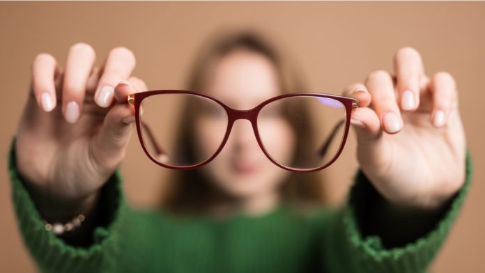 Young girl with poor vision holds glasses with hands forward on beige background.