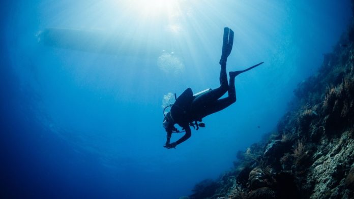 Scuba diver diving towards sun and boat on background underwater