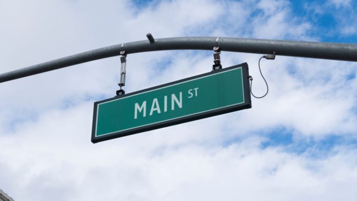 Main Street sign hanging from traffic light, green sign with white lettering and sky with clouds in the background.