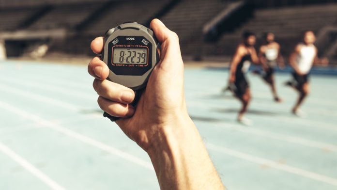Close up of a hand holding a stop watch for time keeping at a running race. Athletes running a race while the time keeper measures time using a stop watch.
