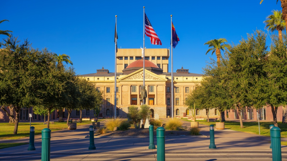 View of the Arizona State Capitol in Phoenix with American, state, and POW flags flying in front of a central statue and landscaped plaza in Phoenix, Arizona.